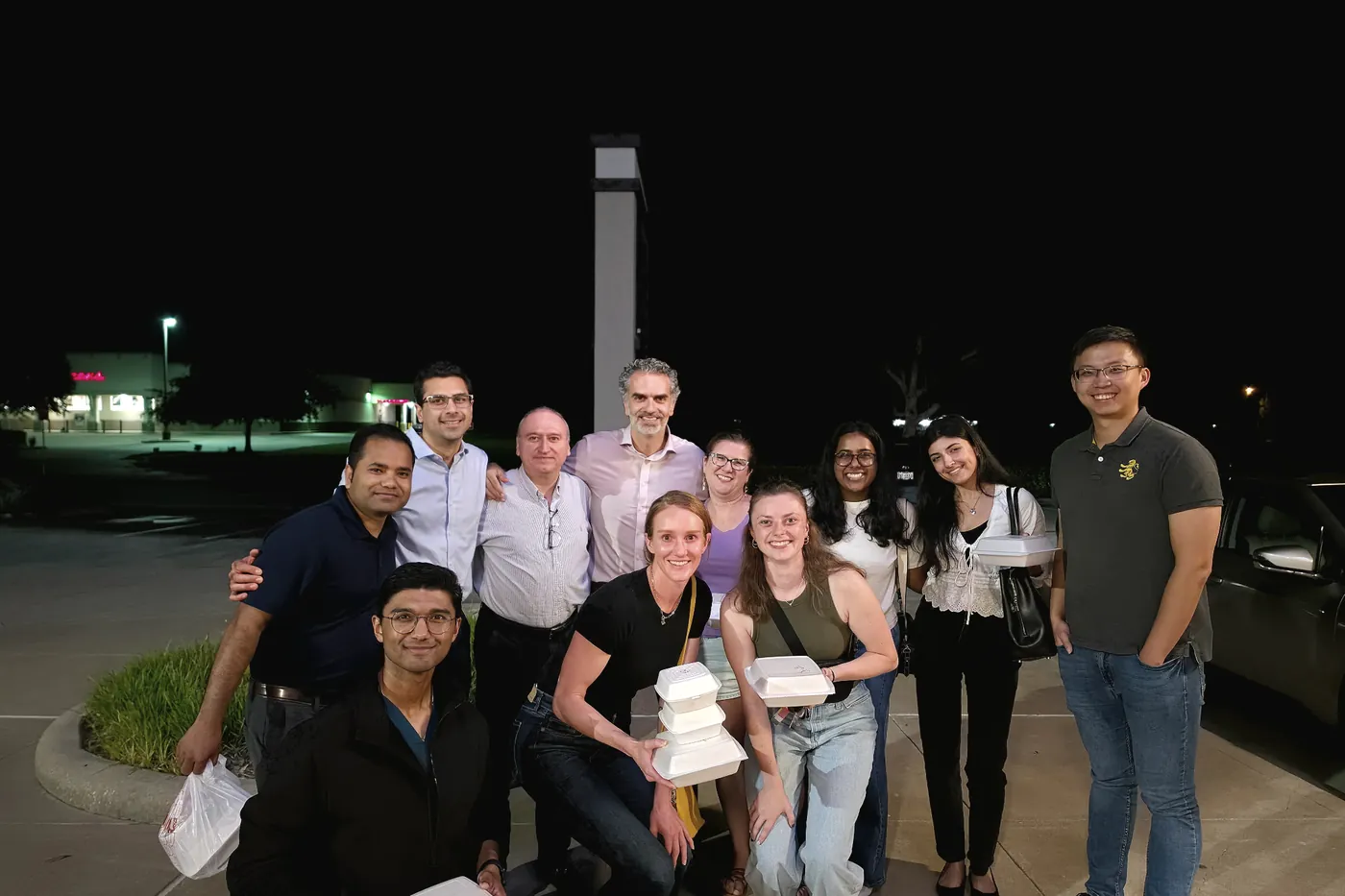 About a dozen lab members standing together outside at night, several holding takeout boxes